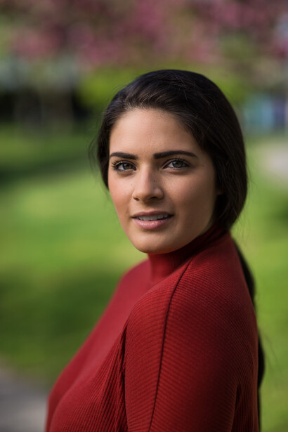 Woman in red cardigan with dark hair looking over shoulder at camera outdoors.