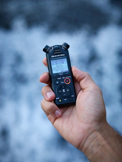 Hand holding a portable audio recording device with small display screen and control buttons against blurred water background.