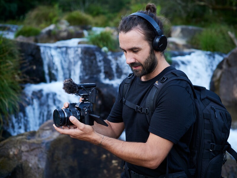 Videographer operating a professional cinema camera near a waterfall, wearing headphones and a backpack.