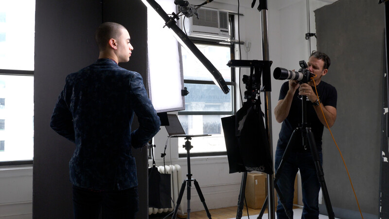 Photographer operating a camera on tripod during a studio portrait session with subject in dark blue jacket.