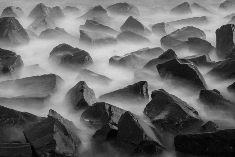 Boulders partially submerged in swirling mist and flowing water, creating an ethereal monochromatic seascape.