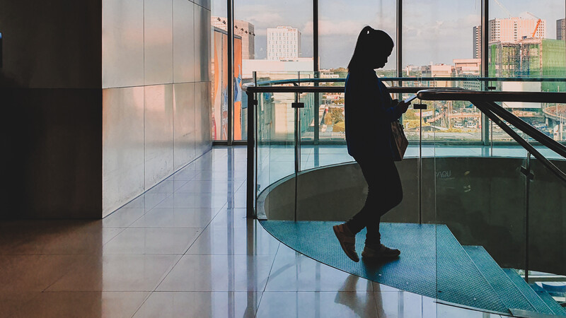 Silhouetted figure in dark clothing standing on a curved blue platform against floor-to-ceiling windows overlooking a city skyline.