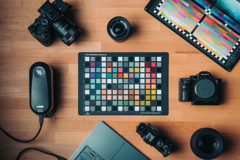 Professional photography gear laid out on wood surface including cameras, lenses, color checker chart, and remote trigger.