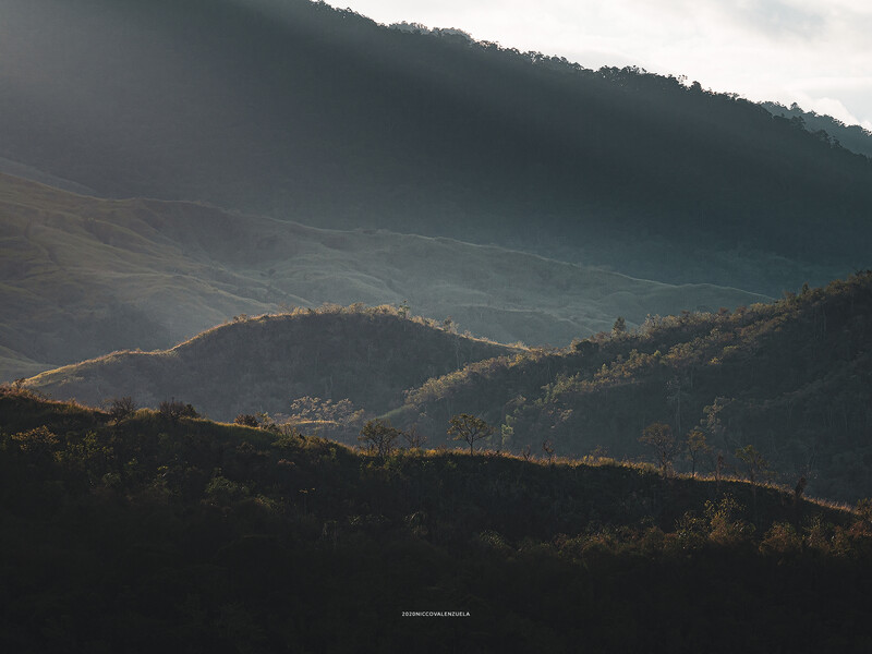 Layered mountain ridges with forested slopes under dramatic directional sunlight and overcast sky.