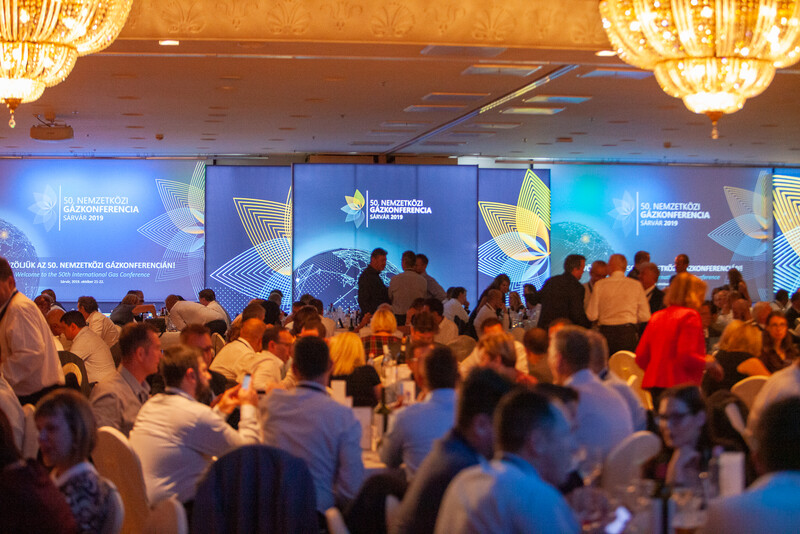 Crowded conference hall with attendees networking beneath illuminated chandeliers and digital presentation screens.