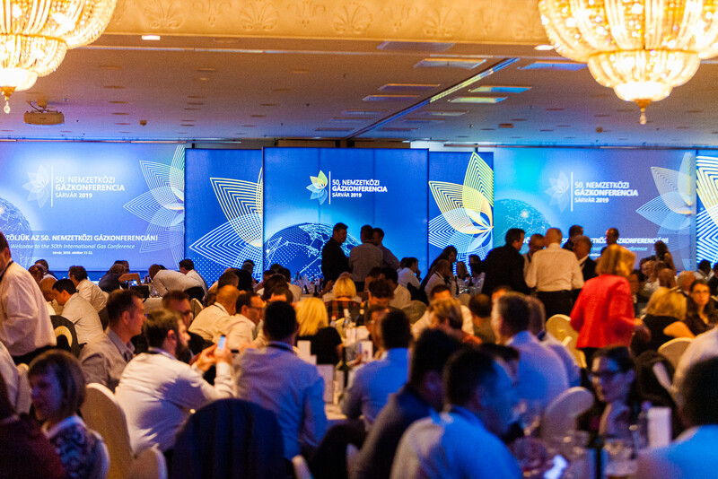 Large conference hall filled with attendees mingling near a blue illuminated stage backdrop.