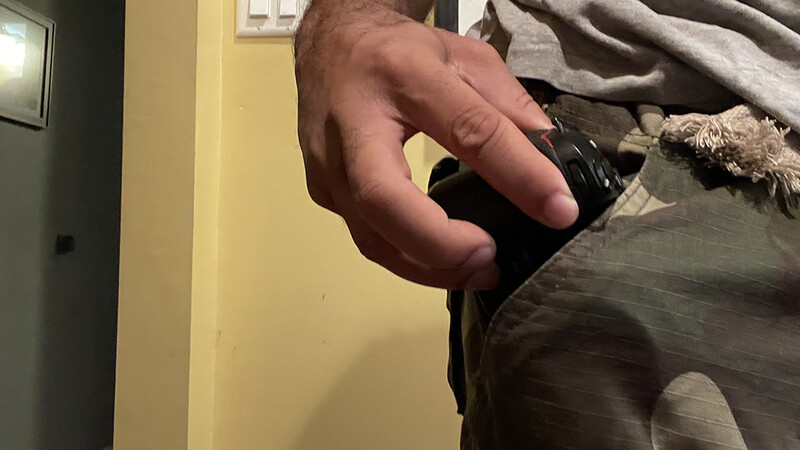 Hand holding a black camera or device against a person wearing gray clothing indoors.