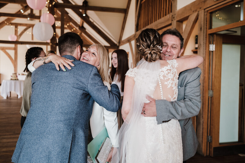 Wedding guests embrace in a group hug inside a rustic venue with exposed wooden beams and white walls.