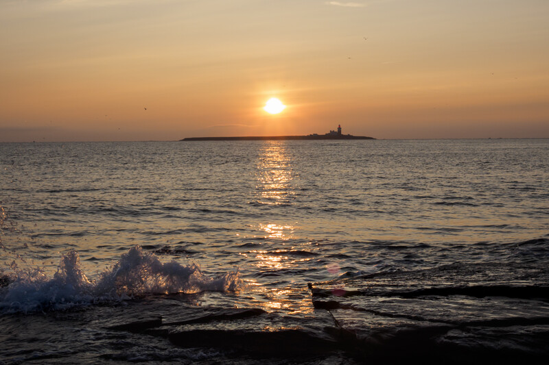 Golden sunset over ocean with waves in foreground and distant lighthouse silhouette.