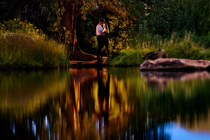 Person standing on a log by a river at golden hour with trees reflected in calm water.