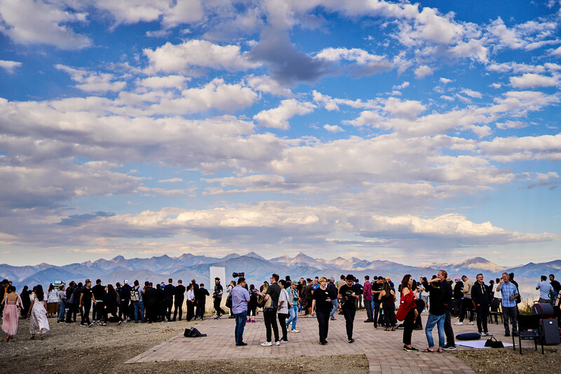 Large crowd of people gathered on a beach with mountains visible in the distant background under a blue sky with white clouds.