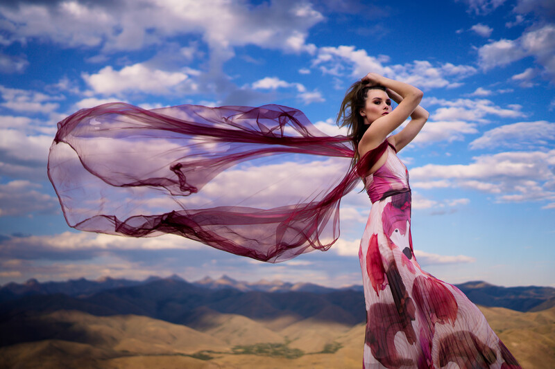 Woman in flowing pink dress with sheer fabric billowing in wind against mountain landscape and blue sky.