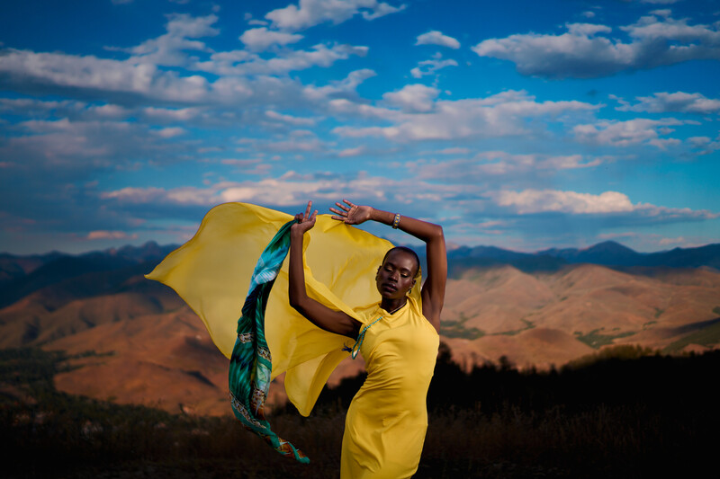 Woman in yellow dress holding flowing yellow fabric against mountain landscape and blue sky.
