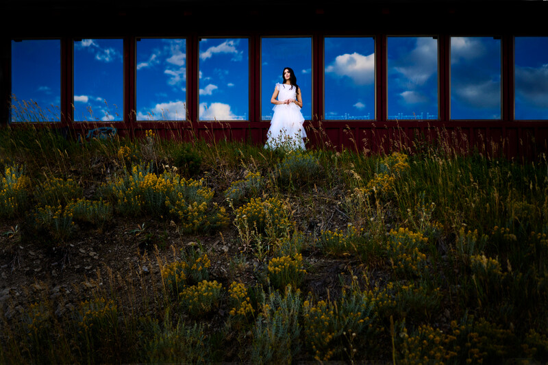 Bride in white wedding dress standing in wildflower field with blue sky windows behind her.