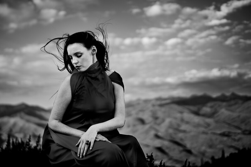 Woman in black sleeveless top seated outdoors with windswept dark hair against cloudy sky and desert landscape.