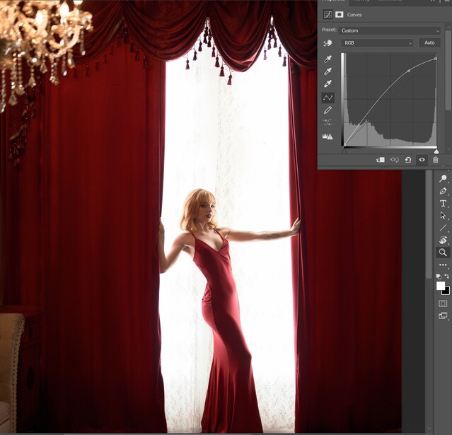 Woman in red gown pulling back heavy burgundy curtains to reveal bright window light.