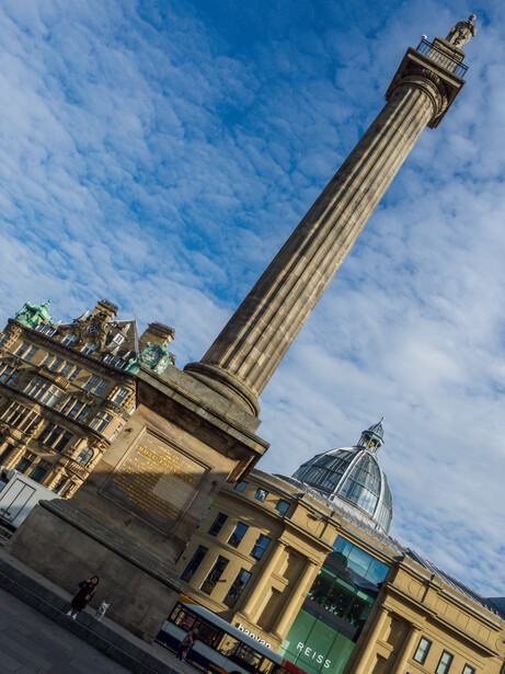 Ornate stone column tower rising against blue sky with classical buildings in foreground.