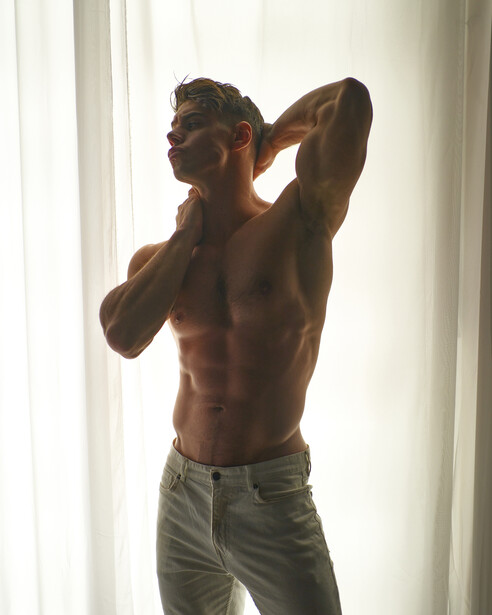 Shirtless male model posing with arms raised behind head near window with natural light.