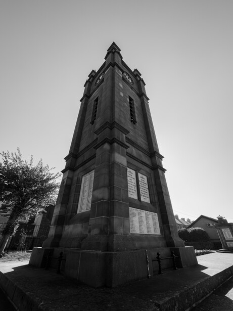 Tall stone tower with peaked roof photographed from below in black and white. The tower features symmetrical window placements and stands against an overcast sky with bare tree branches visible to the left.