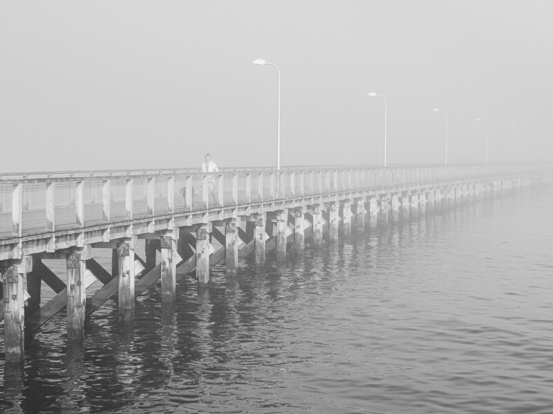 Long pier extending into misty water with repeating support columns and street lamps receding into fog.