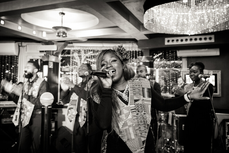 Female singer performing at a microphone with a live band in a glamorous venue with crystal chandeliers.