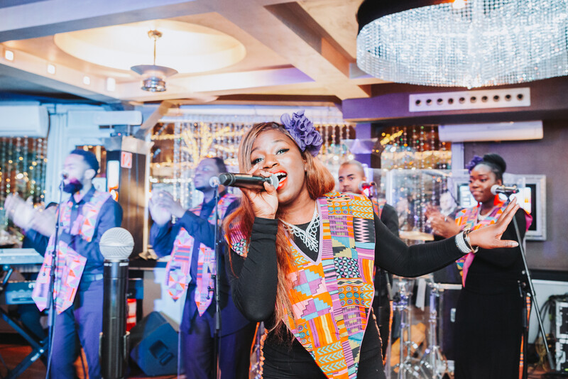 Singer performing at microphone wearing colorful patterned vest in modern venue with purple and gold lighting.