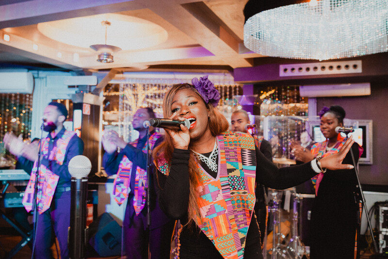 Female vocalist in colorful patterned vest performs at microphone on stage with band members behind her.