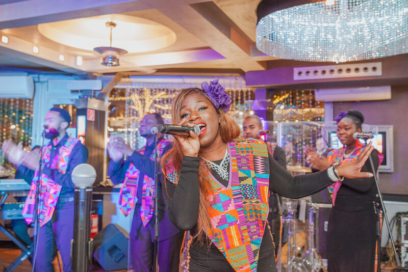 Female singer performing at microphone with band members and colorful stage lighting in venue.