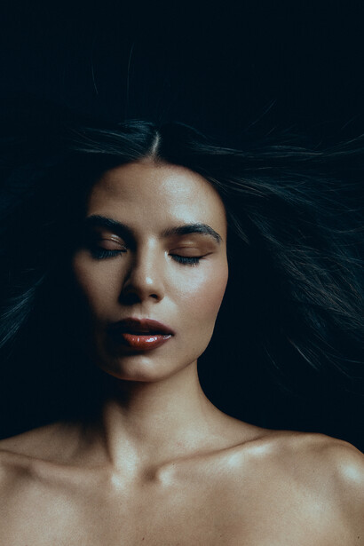 Woman with long dark hair against a black background, eyes closed, with dramatic lighting on her face.