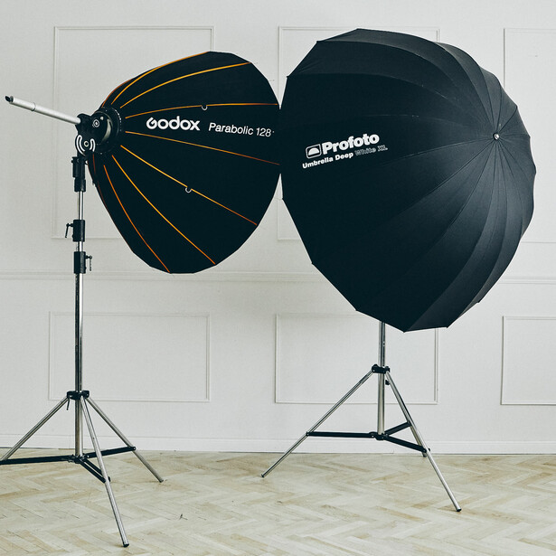 Two large parabolic light modifiers mounted on tripod stands against a white brick wall.