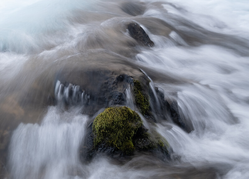 Moss-covered rocks submerged in flowing water with motion blur effect.