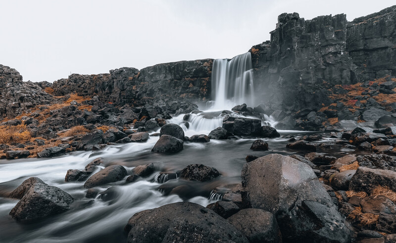 Waterfall cascading over dark basalt cliffs into a misty pool surrounded by moss-covered rocks.