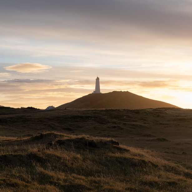 Lighthouse silhouetted against golden sunset sky on a dark volcanic hillside.