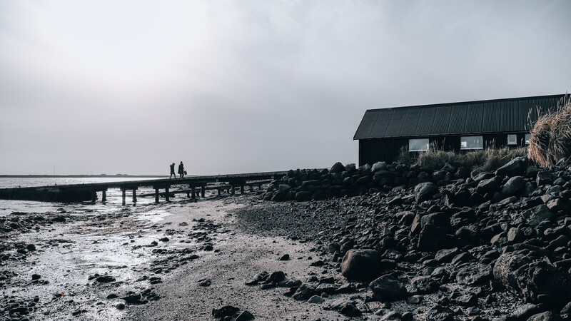 Rocky coastline with weathered boathouse and wooden pier extending into calm water under overcast sky.