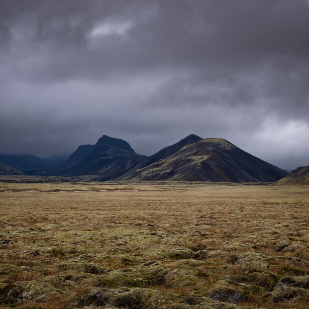 Two dark volcanic mountains rise from a barren, moss-covered plain under dramatic storm clouds.