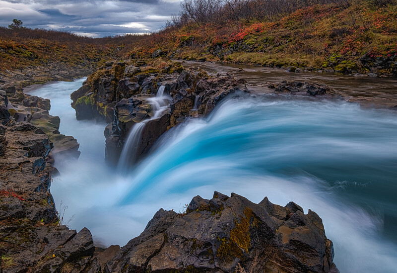 Turquoise waterfall cascading over dark rocky terrain with misty long-exposure effect.