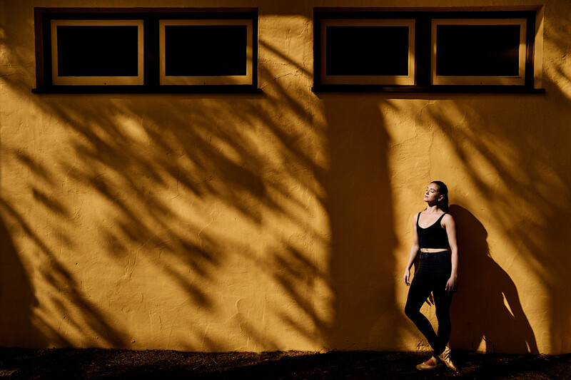 Woman in black athletic wear standing against golden yellow wall with dramatic shadow patterns from window frames.