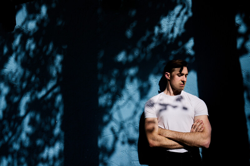 Man in white t-shirt and sunglasses standing with crossed arms against a wall with dappled shadow patterns.