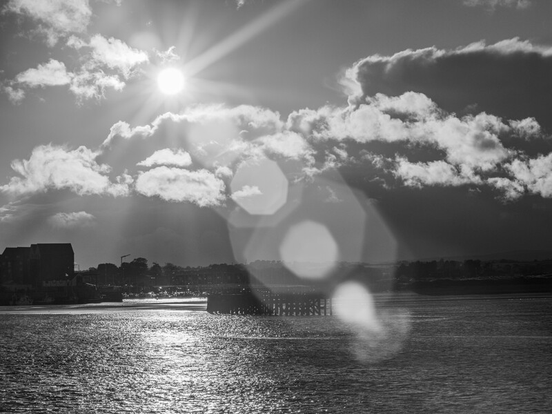 Dramatic black and white landscape of a waterfront with sunburst through layered clouds and lens flare artifacts.