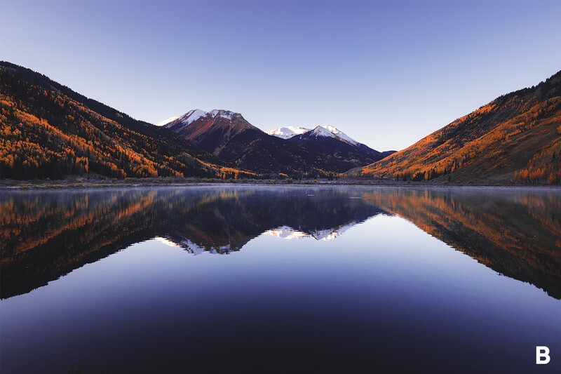 Snow-capped mountain reflected in still alpine lake surrounded by golden larch forests.