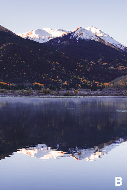 Snow-capped mountain reflected in still alpine lake at dawn, with forested slopes in the foreground.