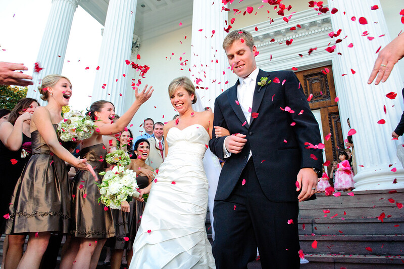 Newlyweds walk down white steps as red rose petals fall around them during a wedding recessional.
