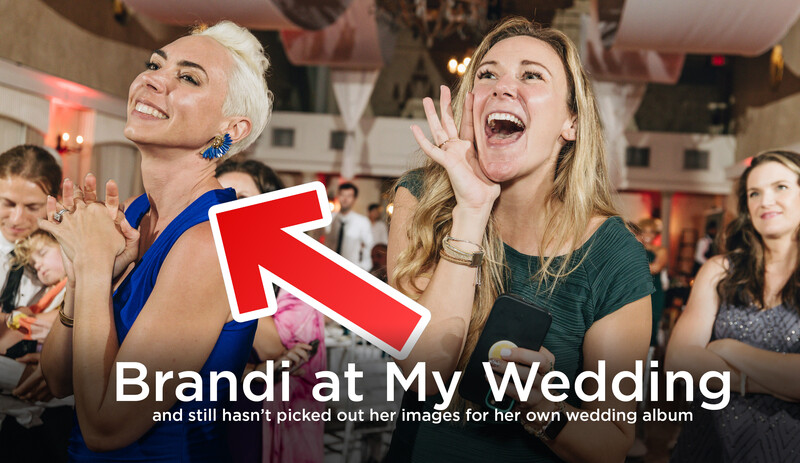 Two women laughing at a wedding reception with guests visible in the background.
