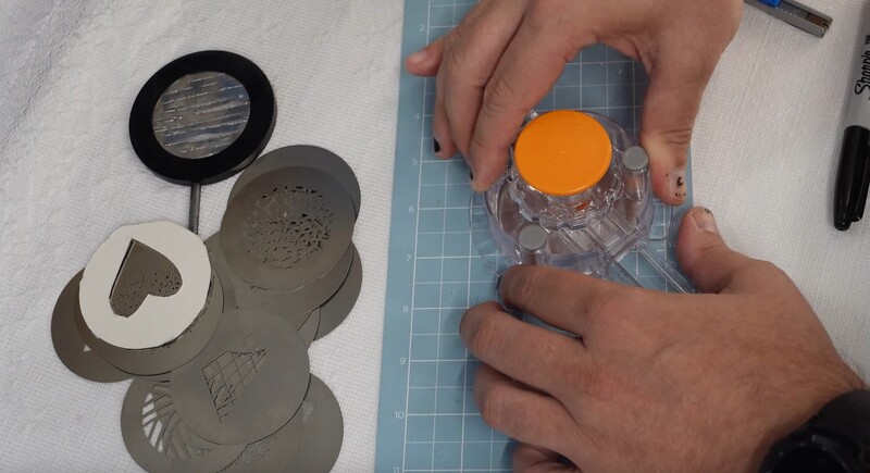 Hands holding a clear container with an orange lid next to stencil templates on a cutting mat.