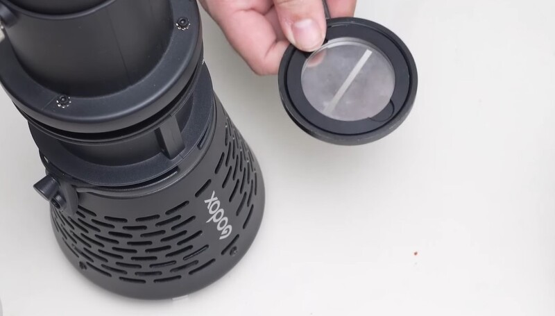 Hand holding a threaded lens cap next to a telephoto lens with ventilation ports.