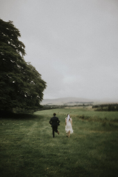 Couple walking through an open field with overcast sky and distant hills.