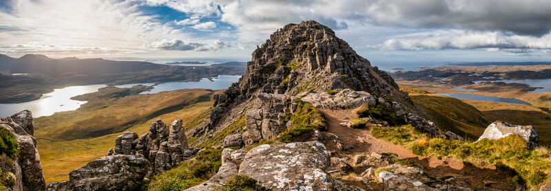 Rocky mountain peak with layered stone formations overlooking multiple lakes and moorland valleys under partly cloudy sky.