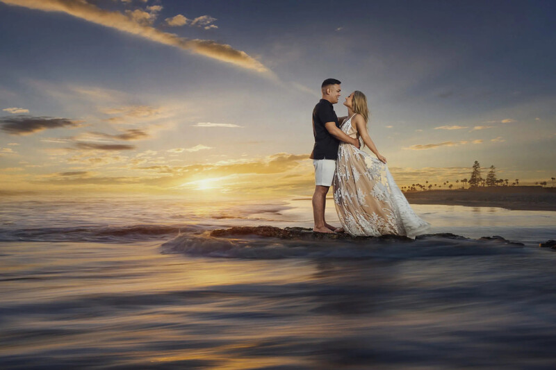 Couple kissing on rocks in shallow ocean water during golden hour sunset.