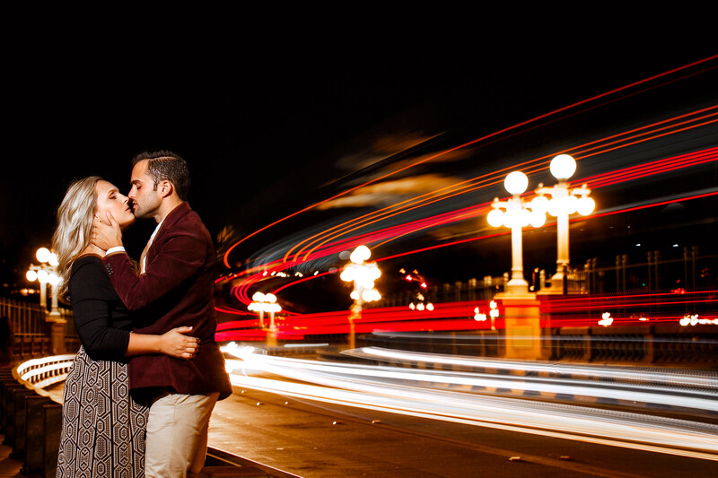 Couple embracing while passing double-decker bus with light trails at night.