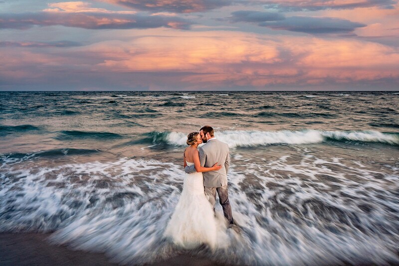Bride and groom standing in ocean waves at sunset with dramatic pink and purple clouds overhead.